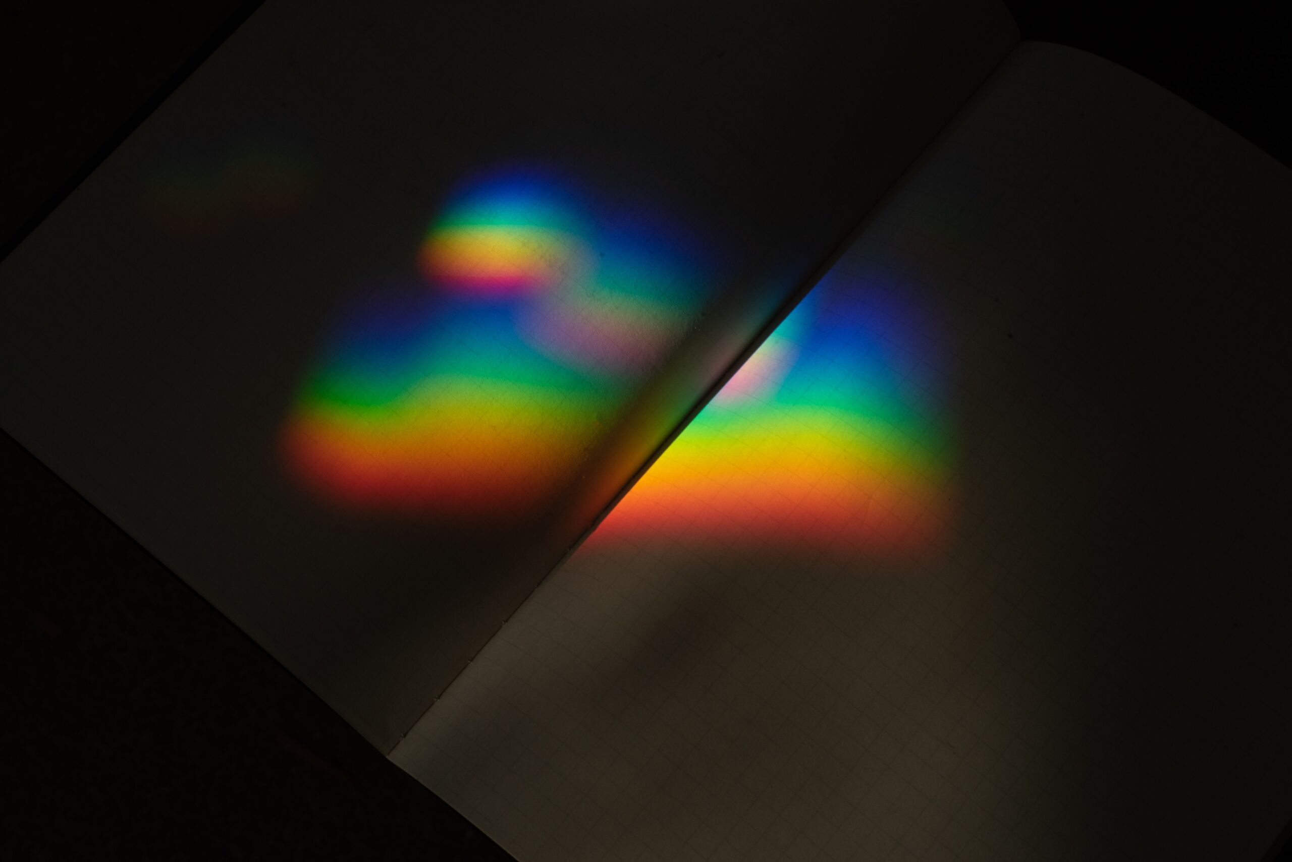 A vibrant prism rainbow reflection on the pages of an open notebook in dark lighting.