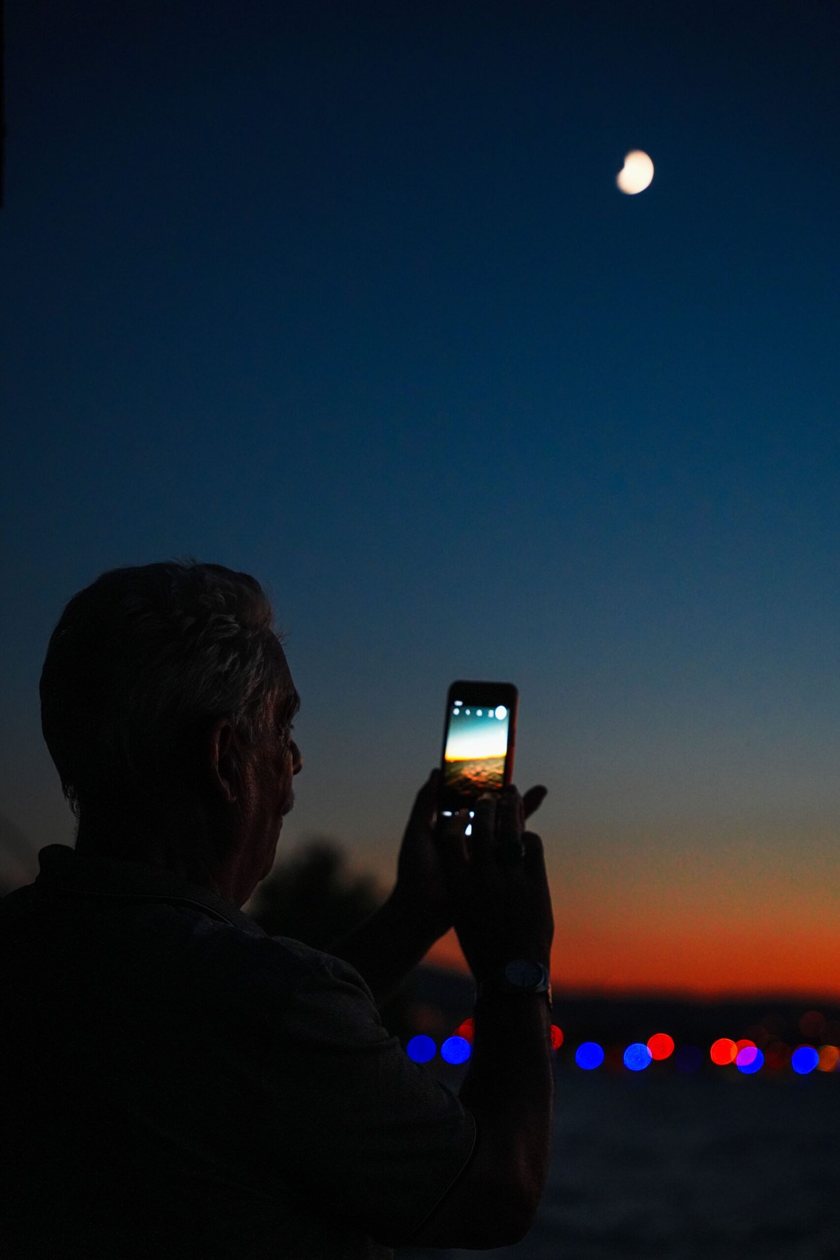 Silhouette of a man taking a photo of the night sky and moon with a smartphone.