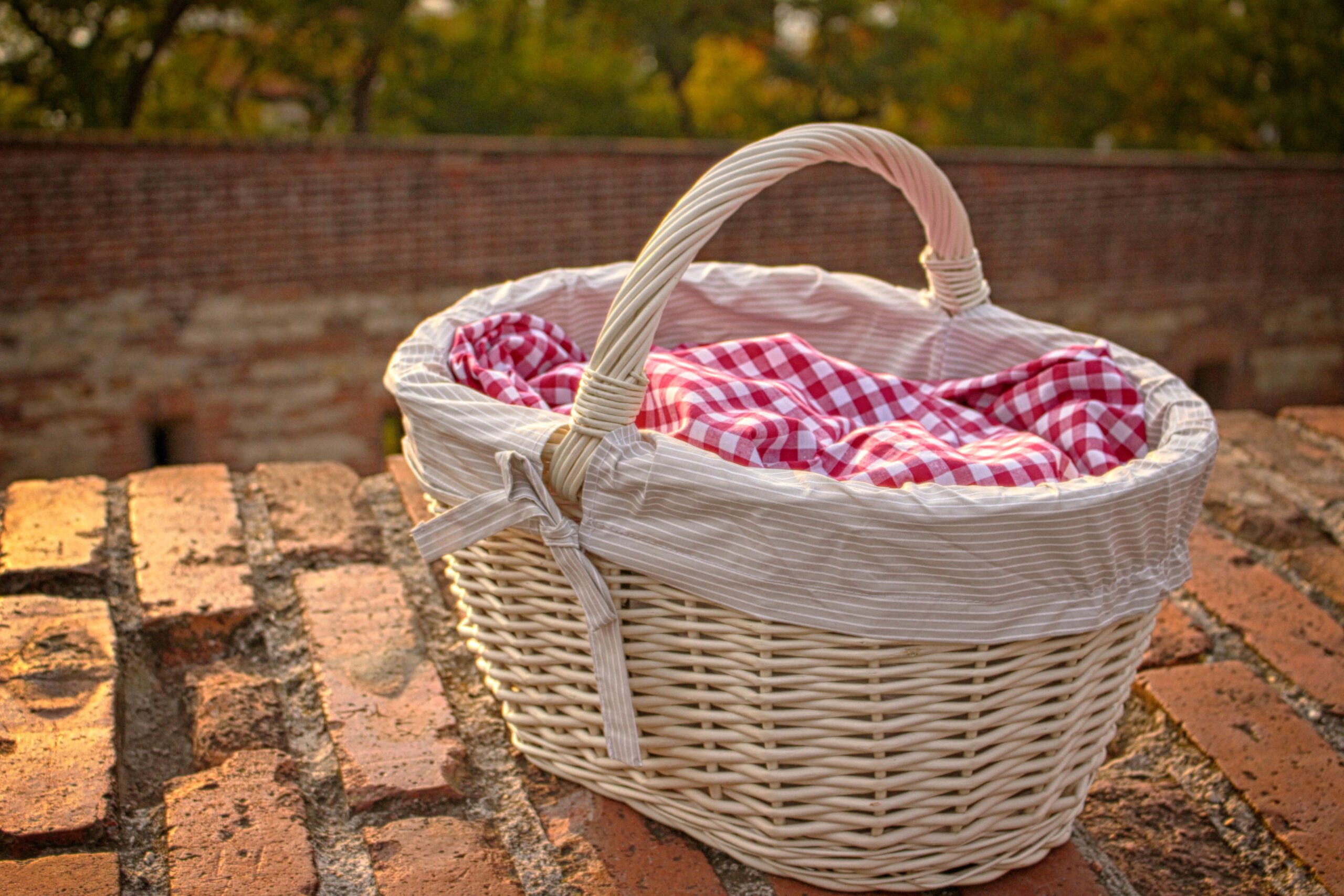 A charming wicker picnic basket with checkered fabric, set against a rustic brick wall outdoors.